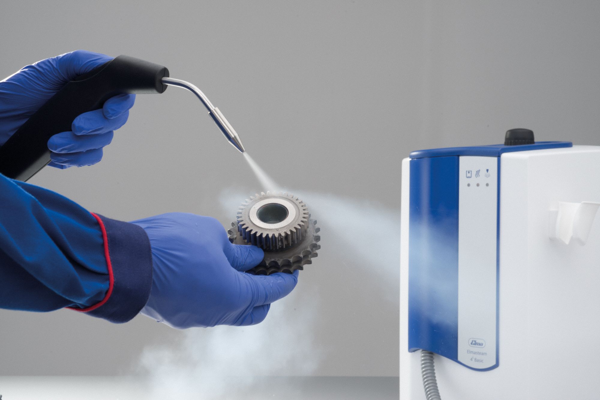 Hands in blue gloves use a steam gun to clean a metal gear next to a white and blue machine.